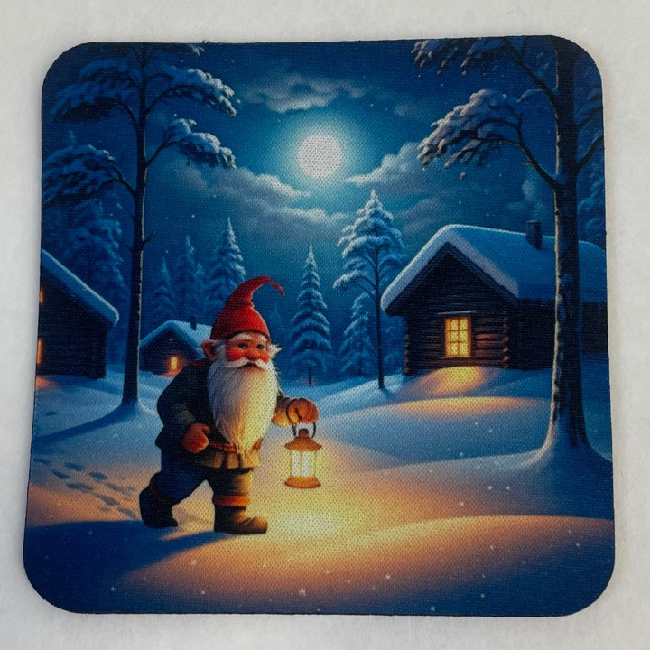 A square coaster with rounded corners featuring a festive winter night scene. The illustration shows a gnome with a red hat and white beard walking through snow while holding a glowing lantern, with snow-covered cabins and trees under a full moon in the background.